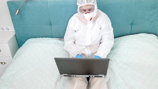 Woman in medical mask and protective suit lying on bed and working on laptop computer. Remote work during global pandemic and covid-19 - 写真・画像