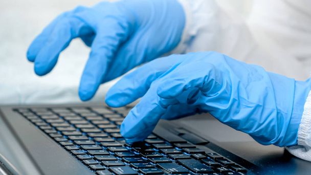 Closeup of fingers in medical gloves typing on laptop keyboard. People working from home and protecting from covid-19 - 写真・画像
