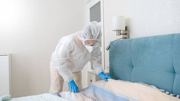 Koruyucu biyo-tehlike kıyafeti içindeki genç ev hanımı covid-19 salgını sırasında yatak hazırlıyor ve kilitleniyor. - Fotoğraf, Görsel