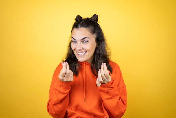 Jonge mooie vrouw dragen sweatshirt over geïsoleerde gele achtergrond doen geld gebaar met de handen, vragen om salaris betaling, miljonair bedrijf - Foto, afbeelding
