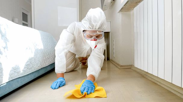 Η νοικοκυρά που φοράει προστατευτική ιατρική στολή πλένει και καθαρίζει το πάτωμα στο σπίτι. Απόσπαση και υγιεινή κατά τη διάρκεια του αποκλεισμού και παραμονή στο σπίτι σε πανδημία - Φωτογραφία, εικόνα