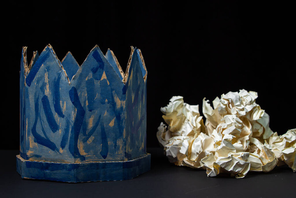 黒の背景に厚紙の王冠。近くには砕いた紙の山がある。紙製品。偽の王冠。ずさんな青い王冠。嘘の力 - 写真・画像