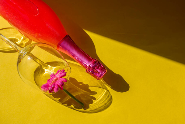 Concepto de fiesta festiva. Luz dura sombras duras. Copa y botella de champán rosa sobre fondo amarillo. La luz del sol revela las formas sólidas de las sombras de vidrio y botellas. Puesta plana. - Foto, imagen