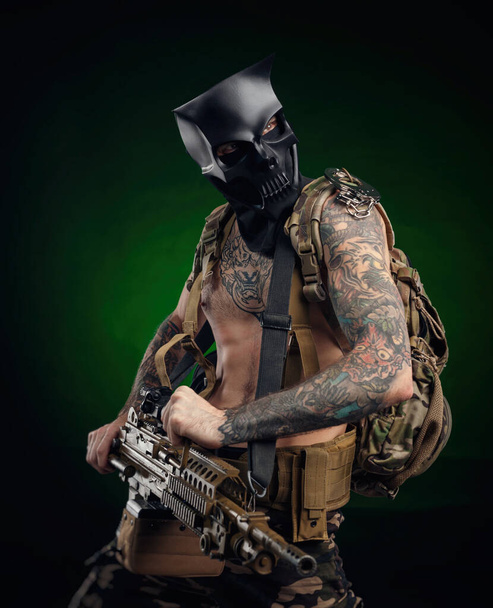 ein Mann mit Totenkopfmaske und einem Maschinengewehr in den Händen mit Tätowierungen - Foto, Bild