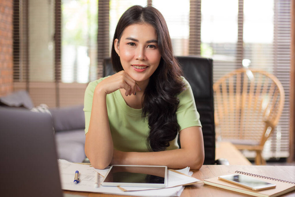 Happy young Asian businesswoman looking staight at camera while sitting and working on her project using her computer and tablet in her living room during work from home - 写真・画像