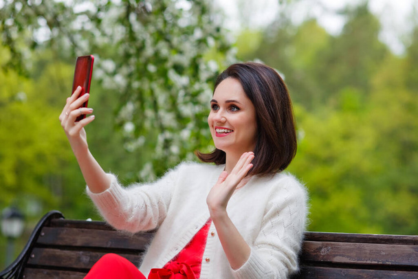 Un mannequin pose avec le sourire dans un parc, assis sur un banc extérieur et parlant sur un smartphone dans ses mains. Portrait d'une jolie fille de beauté brune avec un grand maquillage. - Photo, image