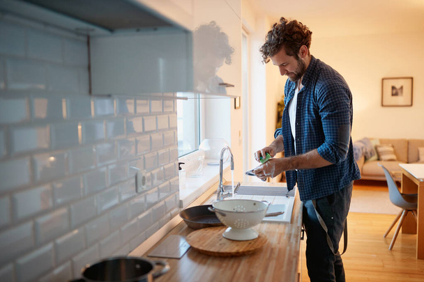 Młody człowiek zmywa naczynia w miłej atmosferze w kuchni. Kuchnia, prace domowe, kwarantanna, domowe - Zdjęcie, obraz