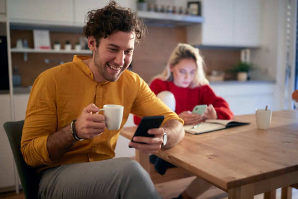 A young couple drinking coffee and surfing the internet on a beautiful morning at the kitchen. Routine, relationship, together - Фото, изображение