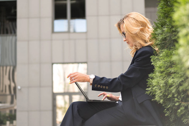 Hermosa mujer rubia en azul marino clásico traje elegante-casual al aire libre cerca del edificio de negocios de alta tecnología. Está trabajando en su portátil. Espacio para texto - Foto, Imagen