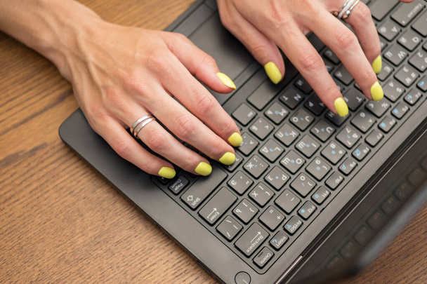 Woman hands on a laptop keyboard on brown table. She working, typing something. Yellow  nail polish on her fingers - Photo, Image