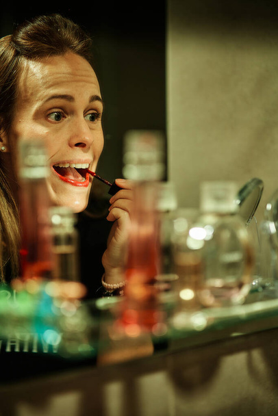 junge Frau, die ihre Lippen zu Hause vor dem Spiegel rot schminkt und zur Seite schaut - Foto, Bild