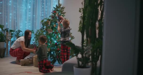 Eine junge Mutter und zwei Jungen im Pyjama schmücken an Silvester den Weihnachtsbaum mit Spielzeug. Vorbereitung auf das neue Jahr und Weihnachten. Das Haus schmücken - Filmmaterial, Video