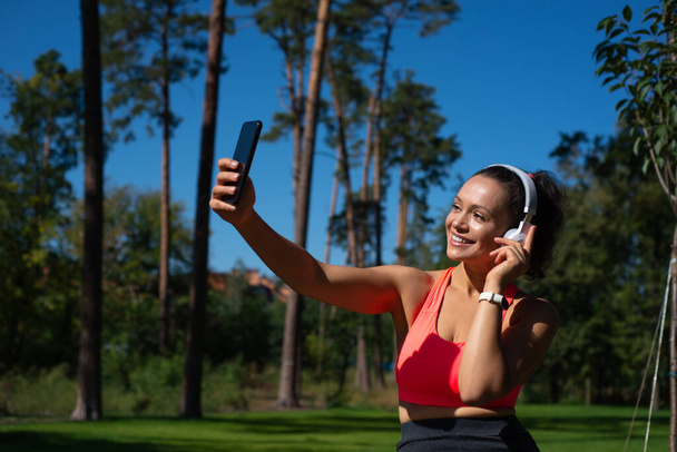 Glimlachende fitte vrouw met hoofdtelefoon maakt selfie tijdens het hebben van een verblijf in het prachtige park. Ontspanning buitenshuis. - Foto, afbeelding