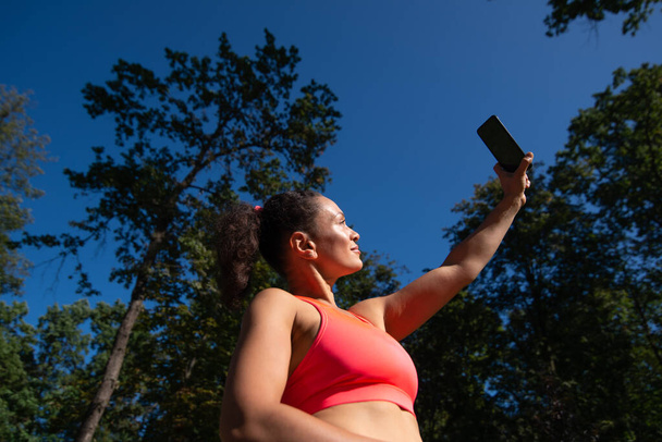 Femme sportive tenant son téléphone portable et faisant du selfie tout en se relaxant après avoir fait du jogging en plein air. Profiter de la belle nature - Photo, image