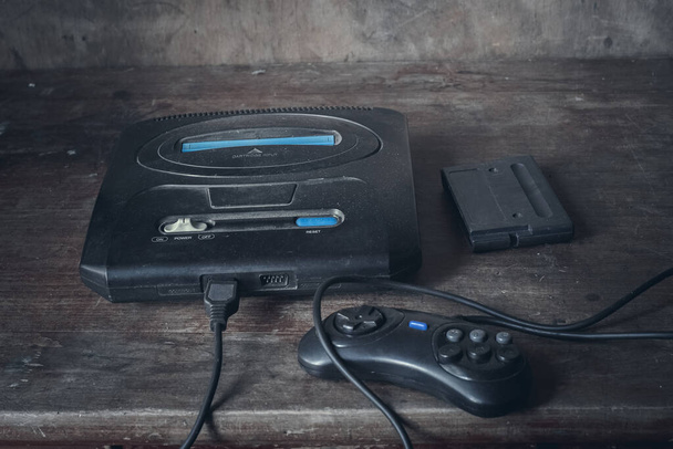 Una vieja consola de juegos en negro, en el polvo sobre una mesa de madera, junto al joystick y el cartucho. - Foto, Imagen