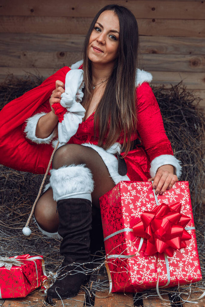 Piękna brunetka dziewczyna w stroju Mikołaja siedzi na słomce z czerwoną torbą prezentów i dekoracje świąteczne - Zdjęcie, obraz