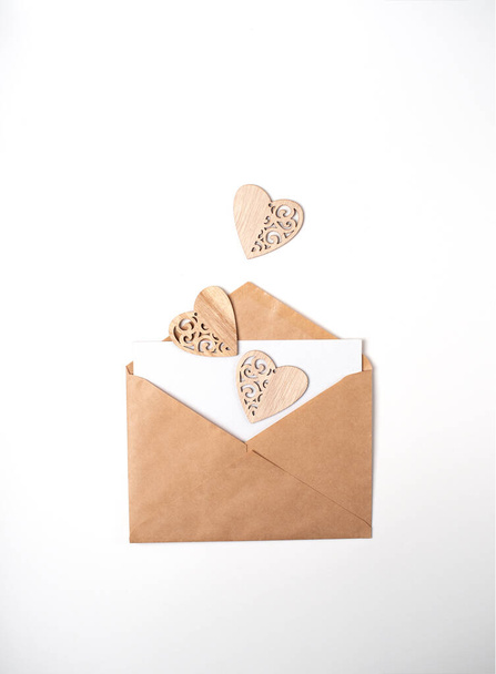 Craft envelope with a blank sheet and three wooden hearts on the light (white) background.. Flat lay, top view. Romantic love letter for Valentine's day concept. - Fotó, kép