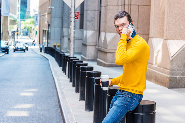 Jonge Latijns-Amerikaan met haarknot, bril op, geel T-shirt met lange mouwen, blauwe jeans, koffie vasthoudend, op straat in New York City, praten op mobiele telefoon, werkpauze. - Foto, afbeelding