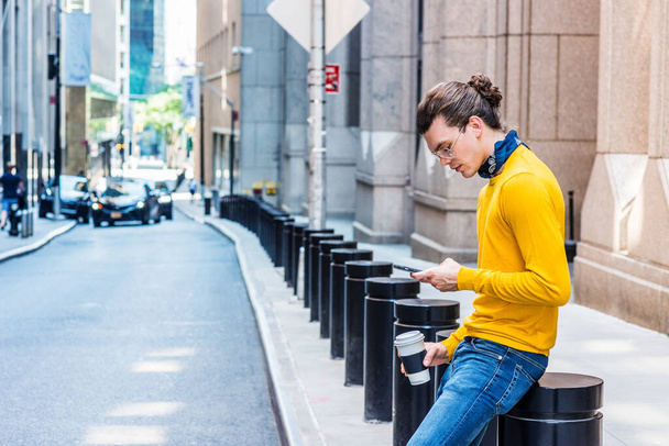 Young Hispanic American with hair bun, wearing glasses, yellow long sleeve T shirt, blue jeans, small black scarf around neck, holding cup of coffee, sitting on street in New York City, texting - Photo, Image