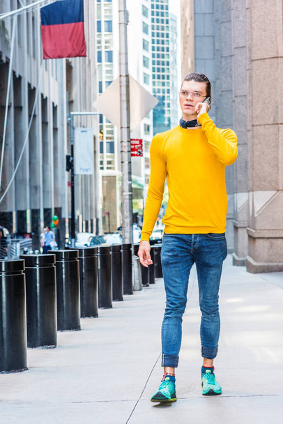 Jonge Latijns-Amerikaan met achterwaarts haar, bril, geel T-shirt met lange mouwen, blauwe jeans, groene sneakers, zwarte sjaal om de nek, lopen op straat in New York City, praten op mobiele telefoon - Foto, afbeelding