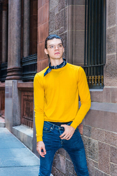 Young Hispanic American Man met haar achterstevoren, met bril, geel T-shirt met lange mouwen, kleine zwarte sjaal rond de nek, staande door oude stijl muur met ramen op straat in New York City - Foto, afbeelding
