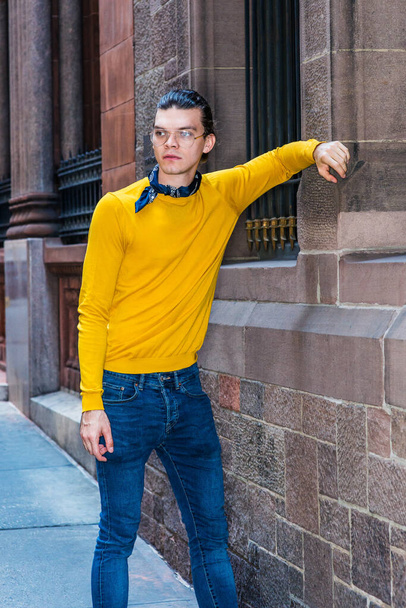 Nuori latino amerikkalainen mies hiukset taaksepäin, silmälasit, keltainen pitkähihainen T-paita, pieni musta huivi kaulan ympärillä, seisomassa vanhan tyylin seinällä ikkunat kadulla New Yorkissa - Valokuva, kuva