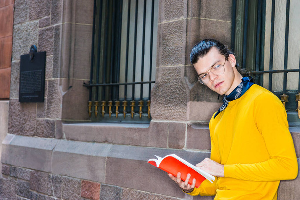 Young Hispanic American Man reading red book outside in New York City, with hair bun, wearing glasses, yellow long sleeve T shirt, small black scarf around neck, standing by wall, looking, thinking - Photo, Image