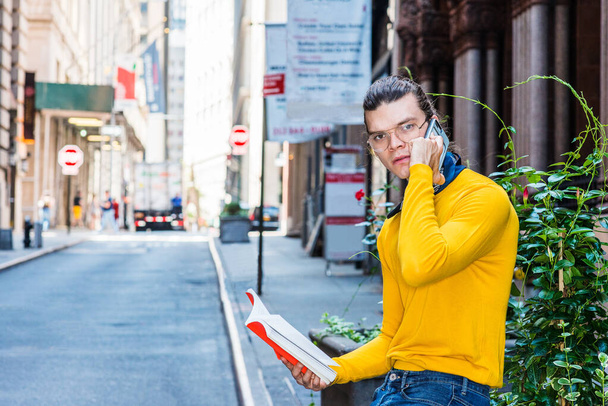 Jonge Latijns-Amerikaanse man met haarknot, bril op, geel T-shirt met lange mouwen, zittend bij groene planten op oude straat in Manhattan, New York. het lezen van rood boek, praten op mobiele telefoon - Foto, afbeelding