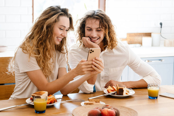 Happy beautiful couple smiling and using cellphone while having breakfast in cozy kitchen - Fotó, kép