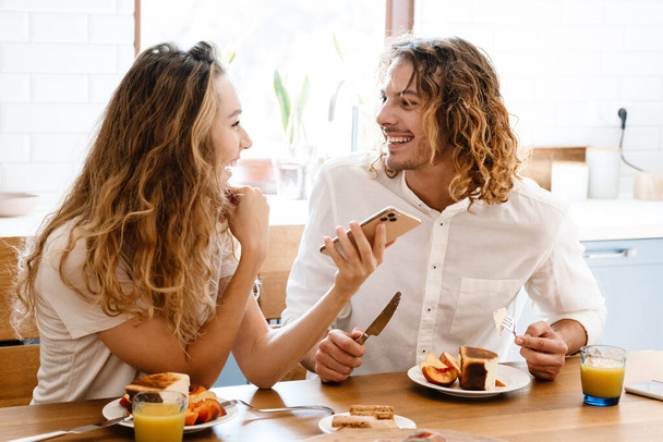 Happy beautiful couple smiling and using cellphone while having breakfast in cozy kitchen - Φωτογραφία, εικόνα