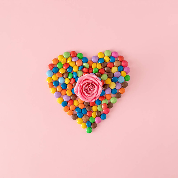 Creative heart shape made with colorful chocolate candy drops with rose in center against pink background. Sweet valentine's day concept - Photo, Image