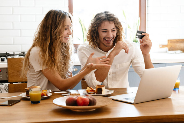 Excited beautiful couple using credit card and laptop while having breakfast in cozy kitchen - Foto, afbeelding