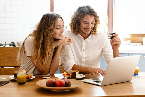 Веселая красивая пара с помощью кредитной карты и ноутбука во время завтрака в уютной кухне - Фото, изображение