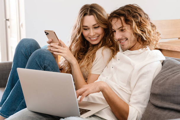 Joyful beautiful couple using cellphone and laptop while resting on couch at home - Фото, зображення