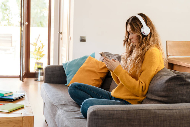 Serious beautiful girl in headphones using cellphone while resting on couch at home - Foto, immagini