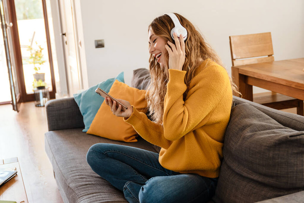 Joyful beautiful girl in headphones using cellphone while resting on couch at home - Фото, изображение