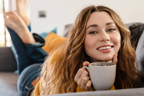 Joyful beautiful girl smiling and drinking tea while lying on couch at home - Fotó, kép