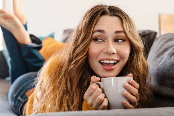 Joyful beautiful girl smiling and drinking tea while lying on couch at home - Фото, зображення