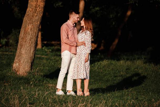 hermosa joven pareja enamorada abrazando y sonriendo en el parque de verano. Hombre con estilo en camisa rosa y mujer en vestido se divierten juntos. historia de amor. espacio de copia. - Foto, Imagen