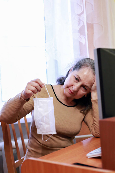 Старшая женщина работает на ноутбуке из дома в хирургической маске из-за ковид-19, активные современные социальные и технологические пожилые люди - Фото, изображение