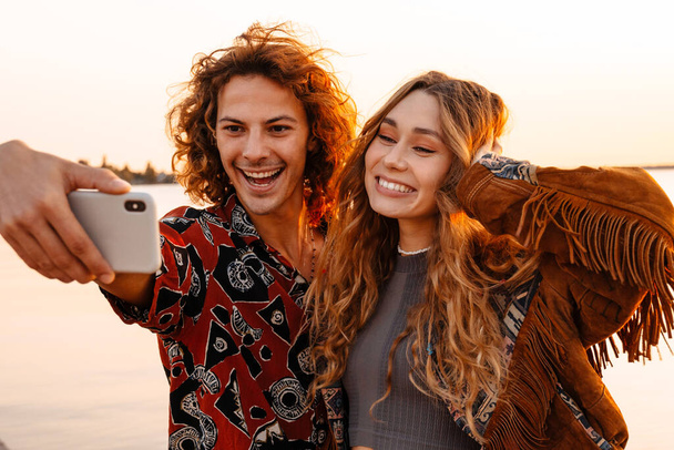 Sahilde cep telefonuyla selfie çeken mutlu, genç bir çiftin fotoğrafı. - Fotoğraf, Görsel
