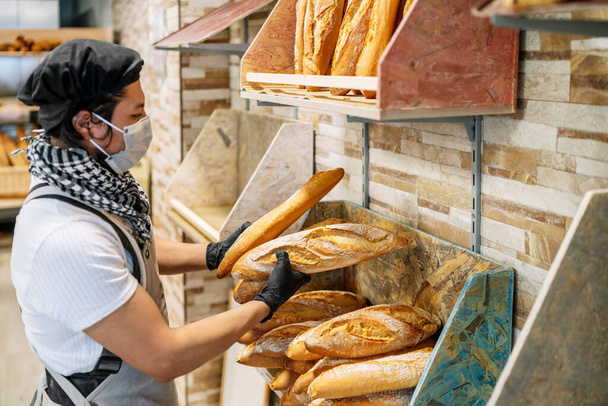 selectieve focus van Latino bakker planken vers gebakken brood met een beschermend gezichtsmasker voor de 2020 covid 19 coronavirus pandemie - Foto, afbeelding