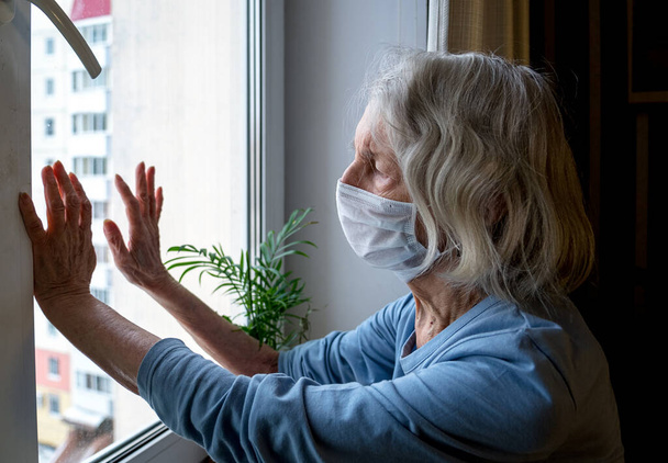Una vecchia donna sola con una maschera medica guarda fuori dalla finestra durante l'isolamento. Una donna depressa a casa durante la pandemia di Covid-19 - Foto, immagini