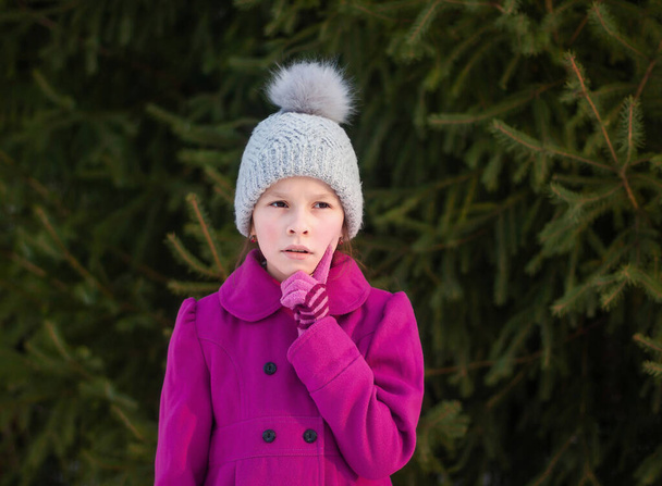 Portrait of thoughtful girl in pink coat and knitted hat on green trees background - Valokuva, kuva