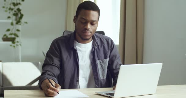 Jovem estudante pensativo afro-americano empresário freelancer senta-se à mesa na frente de laptop trabalhos estudos escreve com caneta sobre papel branco em planos de documentos analisa faz trabalho em papel em casa escritório  - Filmagem, Vídeo