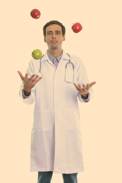 Stúdió lövés fiatal jóképű férfi orvos áll, miközben zsonglőrködik zöld alma és két piros alma - Fotó, kép