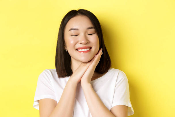 close-up van romantische aziatische vrouw dromen van iets schattig, sluiten ogen en houd handen in de buurt van gezicht, beeldvorming mooi ding, staande over gele achtergrond - Foto, afbeelding
