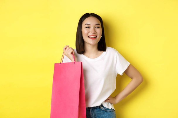 Linda asiática modelo feminino em t-shirt branca, segurando saco de compras e sorrindo satisfeito, em pé sobre fundo amarelo - Foto, Imagem
