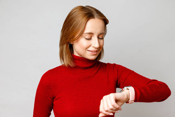 Mulher de negócios elegante feliz em gola alta vermelha olhando para smartwatch em seu pulso, verificando a mensagem na tela, verificando o tempo. Isolado sobre fundo cinzento - Foto, Imagem