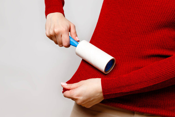 Woman using a lint remover/sticky roller to clean fabrics - red woollen turtleneck from dust, hair, lint and fluff, isolated on grey background. Close up. - 写真・画像
